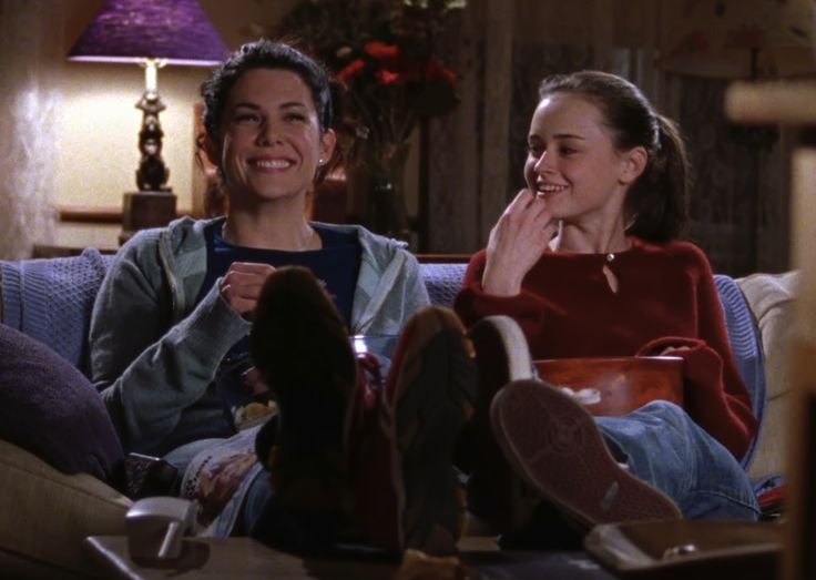 Lorelai and Rory Gilmore sitting on a couch eating popcorn and laughing